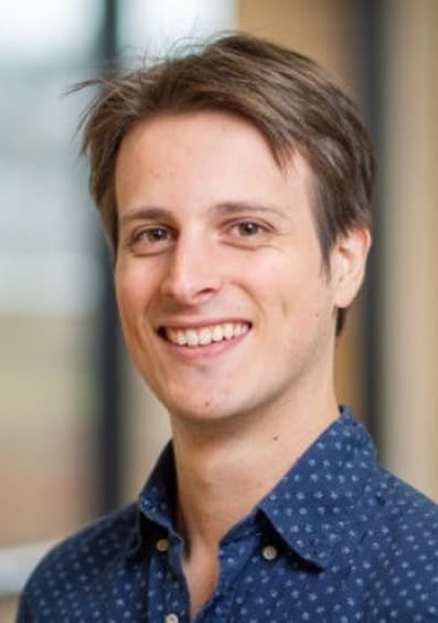 Smiling young man with light brown hair wearing a blue patterned shirt in a softly blurred indoor setting.